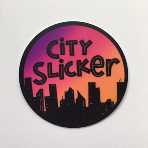 May include: A round sticker with a pink to orange gradient background. The sticker features a black silhouette of a city skyline with the text "city slicker" in black.