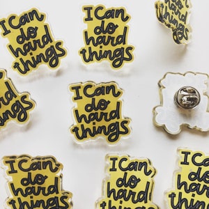 May include: A collection of clear acrylic pins with the phrase "I Can do hard things" in black lettering on a yellow background. The pins have a pin back for attachment. The pins are scattered on a white surface.