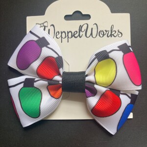May include: A white bow with a black center, featuring a colorful string of lights design. The bow is attached to a white card with the text "Weppet Works" printed on it.