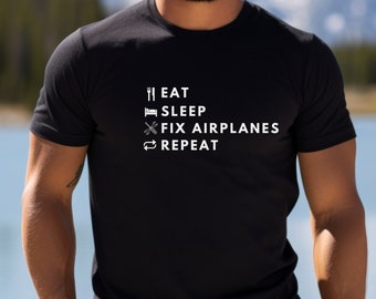 May include: Black crew neck t-shirt with the text "EAT SLEEP FIX AIRPLANES REPEAT" in white. The shirt features icons of a fork and knife, a bed, a wrench and a looping arrow. The background shows a lake and mountains.