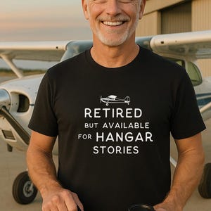 May include: A black t-shirt with the text "RETIRED BUT AVAILABLE FOR HANGAR STORIES" and a small airplane graphic. The man in the image is holding a coffee cup and a black bag, with an airplane in the background.