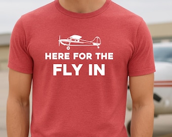 Puede incluir: Camiseta roja con el texto blanco "HERE FOR THE FLY IN". Encima del texto hay un dibujo lineal blanco de un avión pequeño. La camiseta está hecha de un material suave.