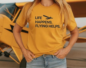 May include: Mustard yellow t-shirt with the text "LIFE HAPPENS. FLYING HELPS." and a small airplane graphic. The shirt is paired with blue jeans. A yellow airplane is in the background.