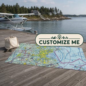 May include: A rectangular mat featuring a detailed aviation sectional chart with yellow, blue, and white navigational markings. A beige canvas tote bag and sunglasses sit nearby on a wooden dock, with a seaplane visible in the water.