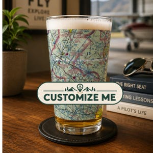 May include: A clear glass pint glass featuring a detailed aviation sectional chart print wrapped around the exterior. The glass contains a golden liquid and sits on a black circular coaster. A white label with green text reads CUSTOMIZE ME.
