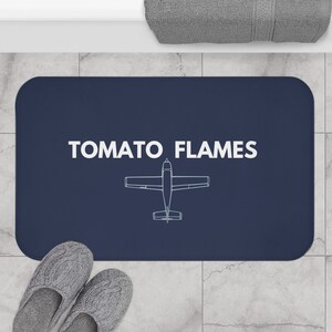 May include: A navy blue bath mat with the text "TOMATO FLAMES" in white and a white outline of a small airplane.