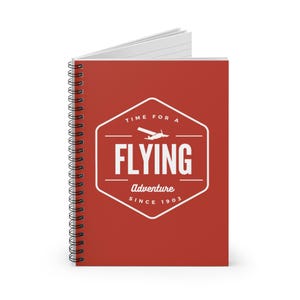 May include: Red spiral notebook with a white hexagonal logo that says "Time for a Flying Adventure Since 1903". The logo features a small airplane silhouette.