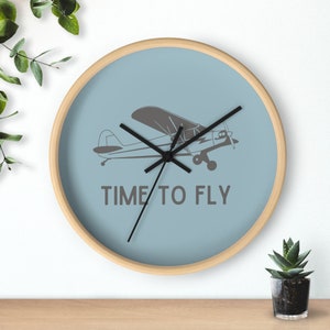 May include: A circular wall clock with a pale blue face and a wooden surround. The clock displays a grey aeroplane graphic and the words "TIME TO FLY". Black clock hands show the time.