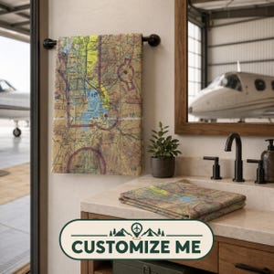 May include: A rectangular towel featuring a detailed aviation sectional chart print with yellow, blue, and brown topographic markings. One towel hangs on a black metal bar, while a second folded towel rests on a bathroom vanity countertop.