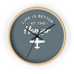 May include: A blue round clock with a wood frame. The clock face has white text that reads "Life is better at the hangar" with a white airplane silhouette. The clock hands are black.