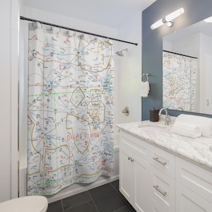 May include: A white shower curtain featuring a detailed map of Dallas-Fort Worth, Texas. The map displays major highways and roads, with labels for cities and towns. The shower curtain is hanging in a bathroom with white tiled walls and a white vanity.