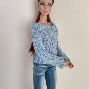 May include: A fashion doll with long auburn hair wearing a light blue long-sleeved knit sweater featuring a cable knit pattern on the chest and ribbed texture on the sleeves and torso, paired with blue denim jeans that have a faded wash finish.