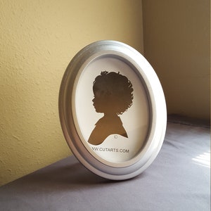 May include: A silver oval picture frame with a silhouette of a child inside. The frame has a raised edge and a website address printed on the bottom: www.cutarts.com
