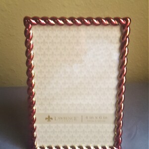 May include: A red and gold picture frame with a rope-like design around the edge. The frame is 4 inches by 6 inches and is made by Lawrence & Co.