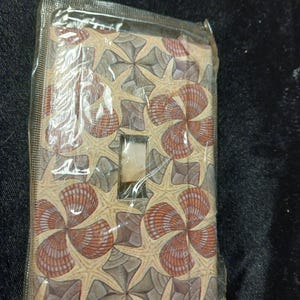 May include: A decorative light switch cover featuring a repeating pattern of starfish and seashells in shades of tan, brown, and gray. The cover is rectangular with a single switch opening and is sealed in clear plastic.