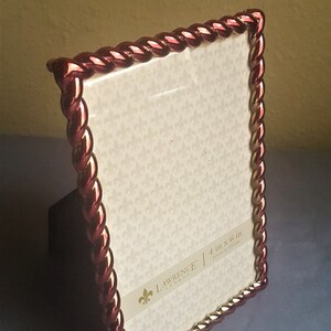 May include: A red and gold picture frame with a rope-like design. The frame is 4 inches by 6 inches and is made by Lawrence Frames.