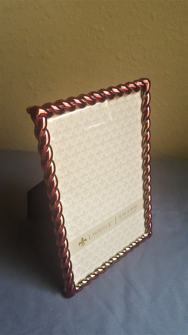 May include: A red and gold picture frame with a rope-like design. The frame is 4 inches by 6 inches and is made by Lawrence Frames.