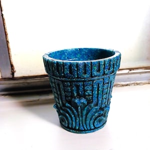May include: A small, blue ceramic planter with intricate, raised designs. The planter has a textured surface and a slightly flared rim.
