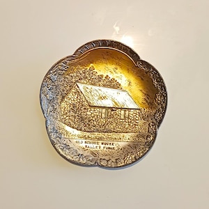 May include: A round, scalloped-edge metal dish with a gold-toned finish. The center features an embossed illustration of a stone building with a gabled roof. Below the building, the text reads: OLD SCHOOL HOUSE VALLEY FORGE PA.