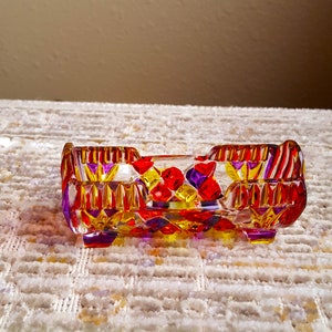 May include: A small, rectangular glass dish with a geometric pattern in shades of red, yellow, and purple. The dish has a slightly raised rim and sits on four small feet.