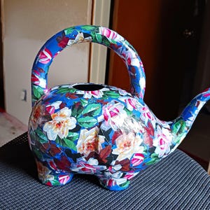 May include: A blue watering can with a floral design. The can is shaped like a teapot with a spout and a handle. The flowers are painted in shades of pink, white, and green.