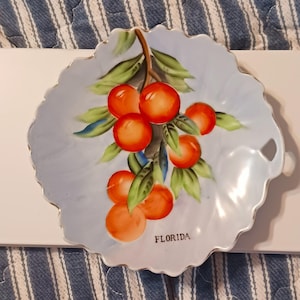 May include: A light blue ceramic dish with a scalloped edge and a painted design of orange citrus fruits on green leafy branches. The word FLORIDA is printed in black capital letters near the bottom of the dish, which features a small side handle.