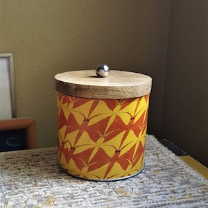 May include: A yellow and orange cylindrical container with a wooden lid and a silver knob. The container has a geometric pattern of overlapping shapes.