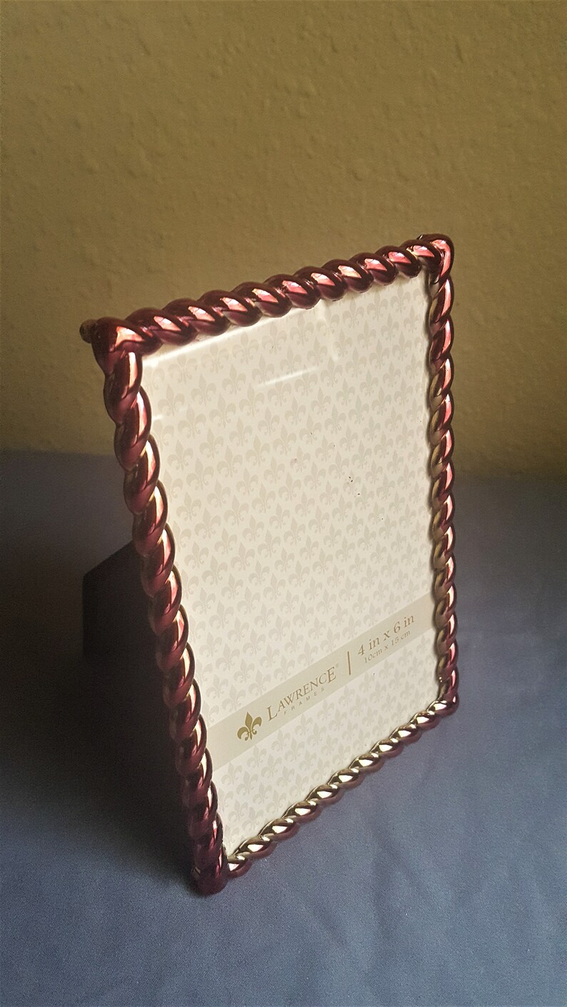 May include: A red and gold picture frame with a rope-like design. The frame is 4 inches by 6 inches and is made by Lawrence Frames.