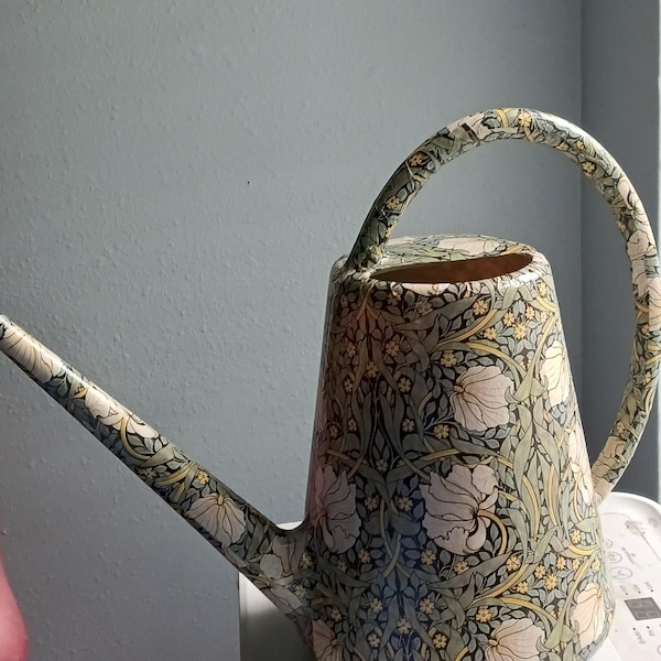 Pode incluir: Um regador de metal com um padrão botânico detalhado de flores brancas, detalhes amarelos e folhagem verde-sálvia sobre um fundo escuro. O recipiente inclui um bico longo e fino, uma alça curva e uma abertura superior para enchimento.