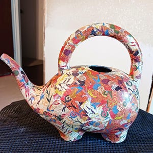 May include: A colorful watering can shaped like an elephant with a floral and bird pattern. The watering can is made of plastic and has a handle on the top.