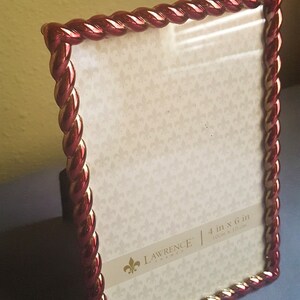 May include: A red and gold picture frame with a rope-like design. The frame is 4 inches by 6 inches and is made by Lawrence Frames.