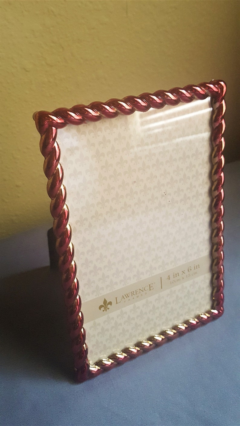 May include: A red and gold picture frame with a rope-like design. The frame is 4 inches by 6 inches and is made by Lawrence Frames.
