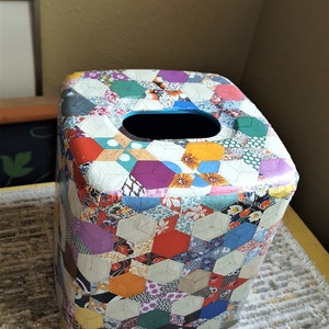 May include: A square tissue box cover with a patchwork design featuring various colors and patterns. The cover is made of a material that resembles fabric and has a blue opening for tissues.