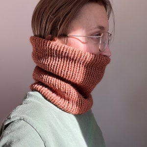 May include: A person wearing a brown knitted neck warmer and glasses. The neck warmer is made of a soft, textured yarn.