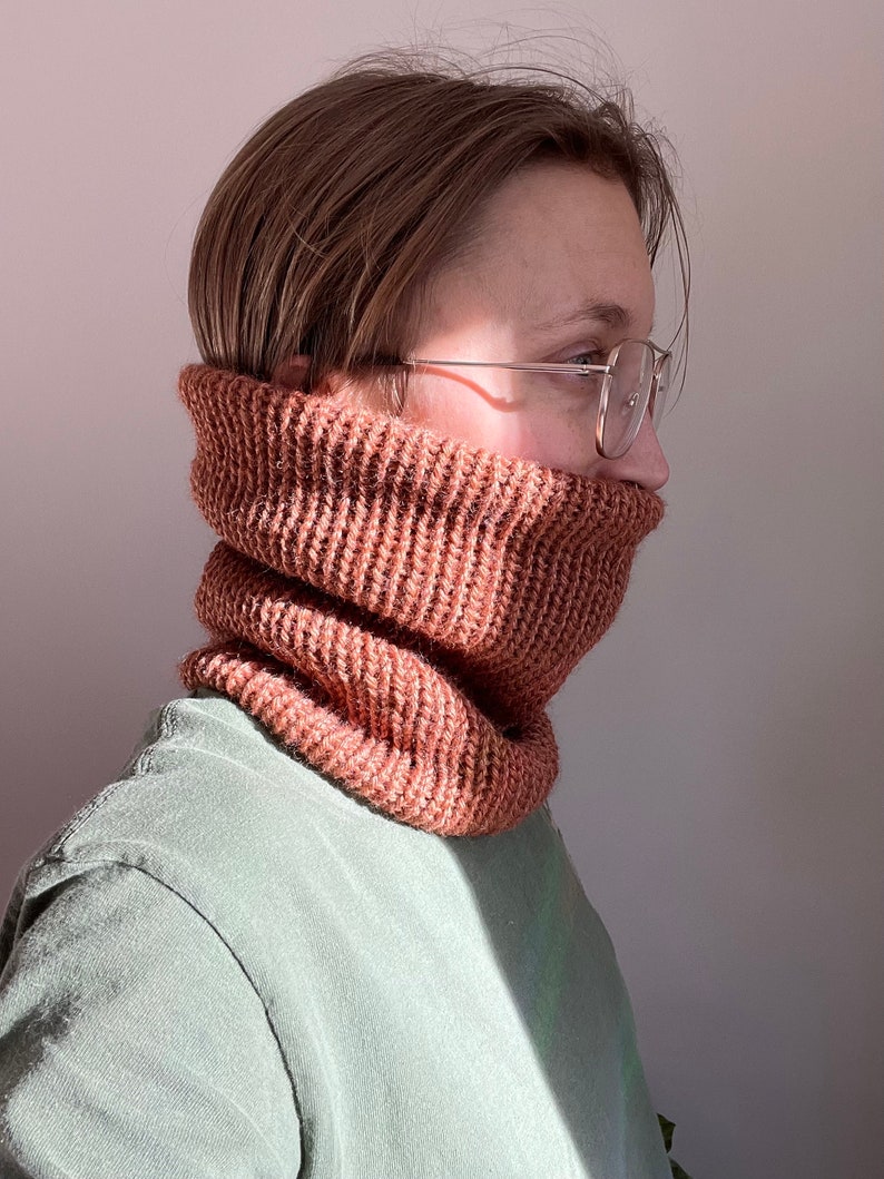 May include: A person wearing a brown knitted neck warmer and glasses. The neck warmer is made of a soft, textured yarn.