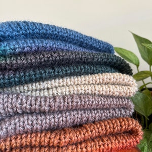 May include: A stack of six crocheted scarves in various shades of blue, green, beige, brown, and orange. The scarves are folded and arranged on a wooden surface.