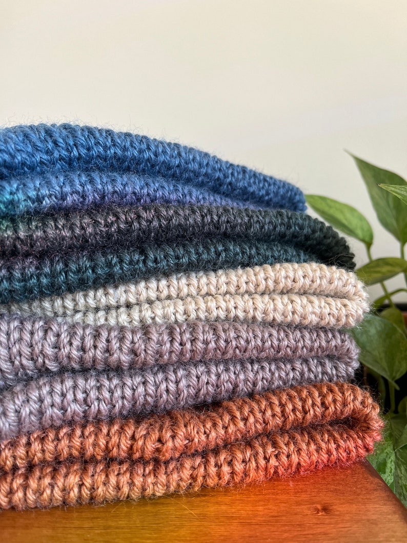 May include: A stack of six crocheted scarves in various shades of blue, green, beige, brown, and orange. The scarves are folded and arranged on a wooden surface.