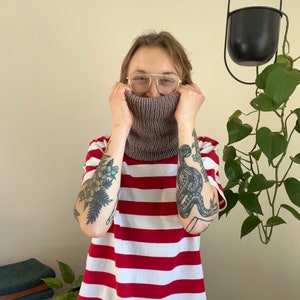 May include: A person wearing a red and white striped shirt with a brown knitted neck warmer. The person has tattoos on both arms.