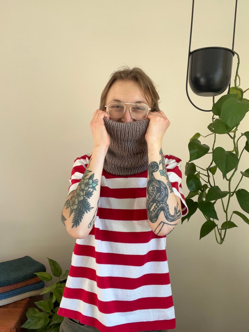 May include: A person wearing a red and white striped shirt with a brown knitted neck warmer. The person has tattoos on both arms.