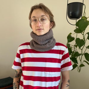 May include: A person wearing a red and white striped shirt with a brown knit neck warmer. The person is wearing glasses and has a tattoo on their arm.