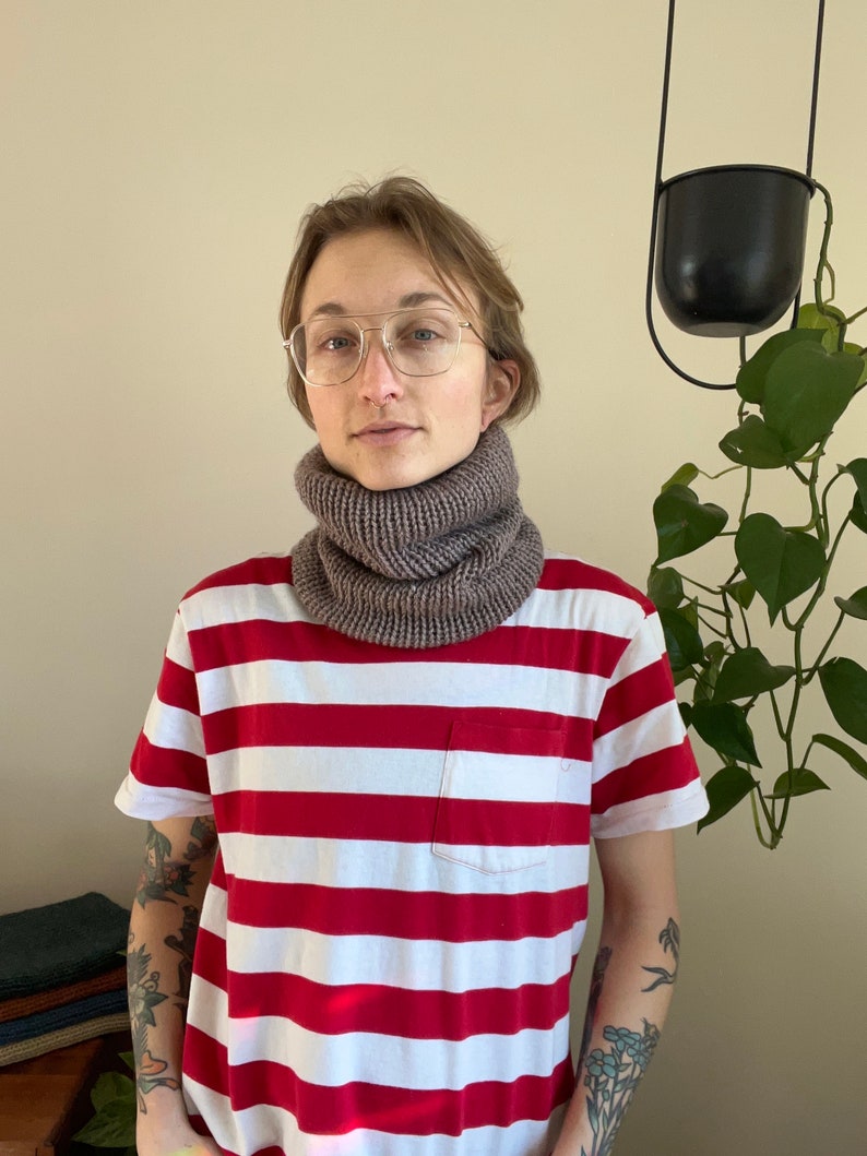 May include: A person wearing a red and white striped shirt with a brown knit neck warmer. The person is wearing glasses and has a tattoo on their arm.