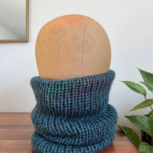 May include: A dark blue and green knitted neck warmer or cowl. The neck warmer is folded over and is sitting on a wooden surface.