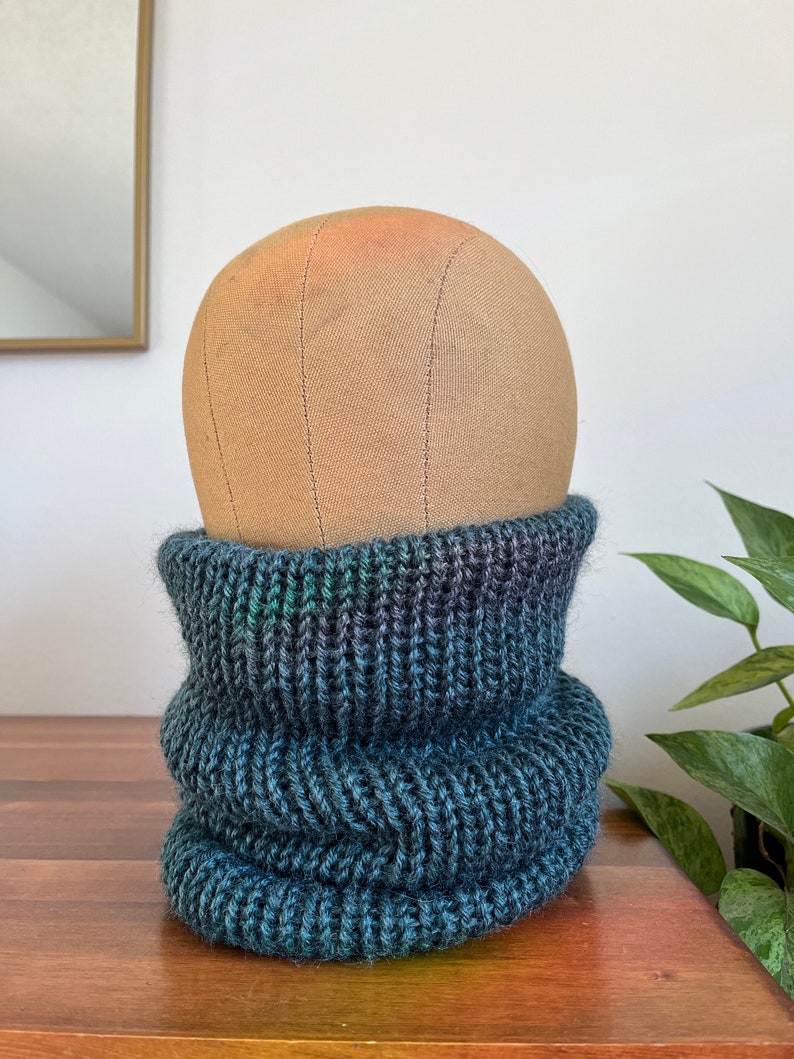 May include: A dark blue and green knitted neck warmer or cowl. The neck warmer is folded over and is sitting on a wooden surface.