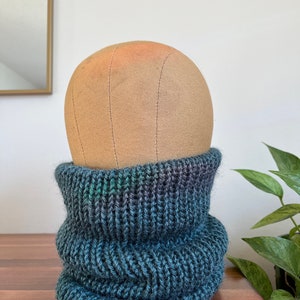 May include: A dark green and black knit neck warmer. The neck warmer is folded over and is sitting on a wooden surface.