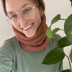 May include: A person wearing a light green long-sleeved shirt with a brown knitted neck warmer. The person is wearing glasses and has a nose ring.