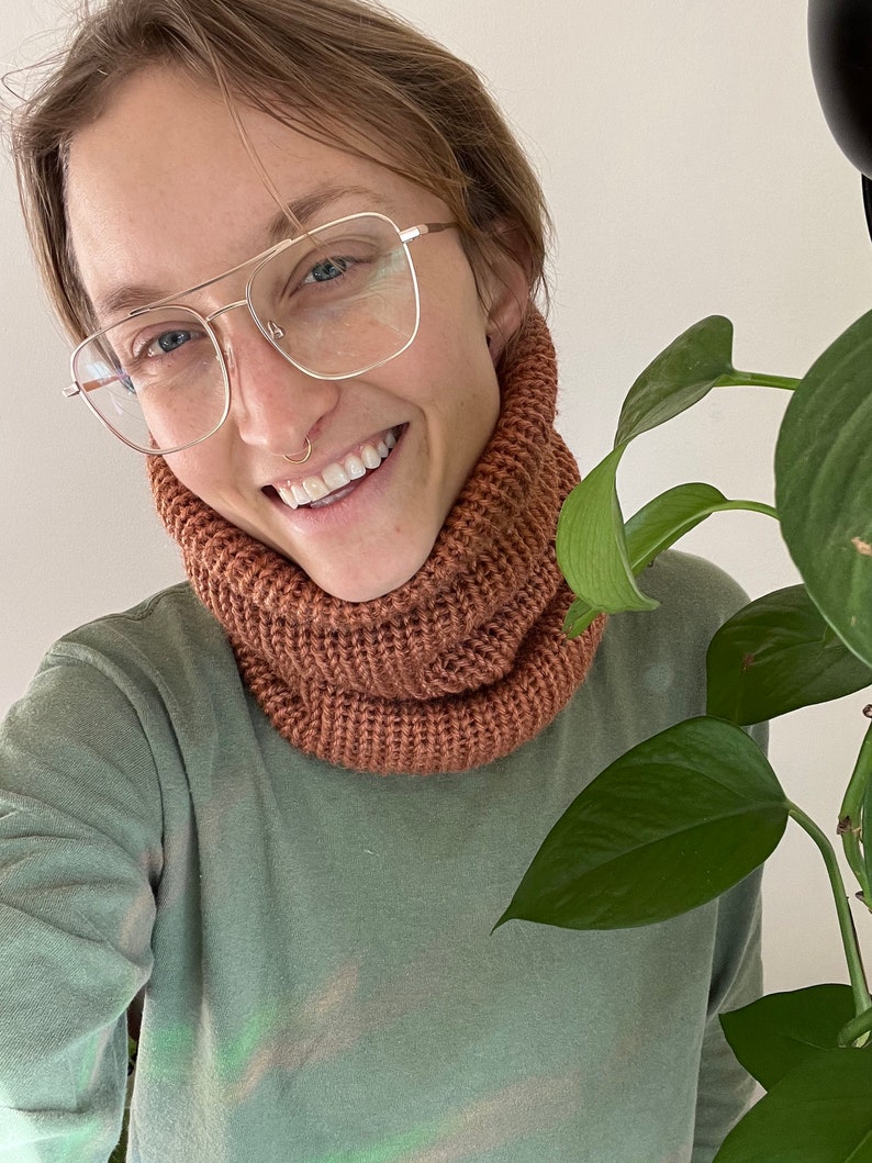 May include: A person wearing a light green long-sleeved shirt with a brown knitted neck warmer. The person is wearing glasses and has a nose ring.