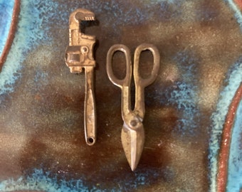 May include: Two miniature tools, a wrench and a pair of scissors, are displayed on a blue and brown patterned surface. The tools are made of metal and have a rustic, antique appearance.