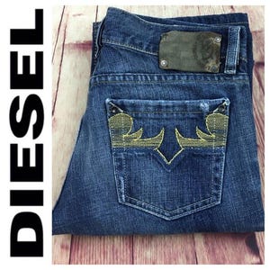 Può includere: Jeans blu in denim piegati. La tasca posteriore presenta un design ricamato color oro. È visibile una toppa in pelle con il nome del marchio. La parola "DIESEL" è stampata in nero a sinistra.