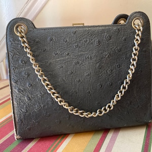 Puede incluir: Un bolso de cuero de avestruz negro con una correa de cadena dorada. El bolso tiene un cierre de broche y una superficie texturizada.