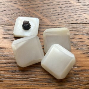 May include: Four white square buttons with a slightly iridescent finish. One button has a small black metal loop on the top.